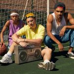 Three men in retro fashion posing with a boombox on a sports court.