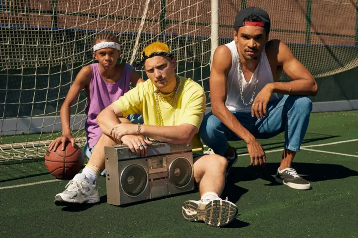 Three men in retro fashion posing with a boombox on a sports court.