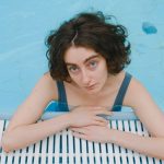 A woman with curly hair leaning on the side of a swimming pool, looking up.