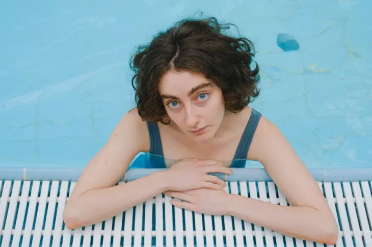 A woman with curly hair leaning on the side of a swimming pool, looking up.