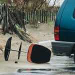A surfboard lying beside a car in a wet, sandy beach area during rainy weather.