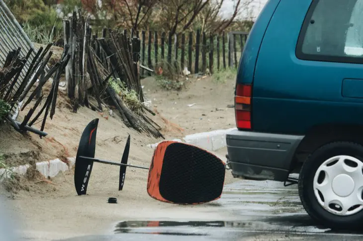 A surfboard lying beside a car in a wet, sandy beach area during rainy weather.