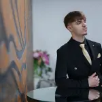 Man in a black suit with a gold tie poses.